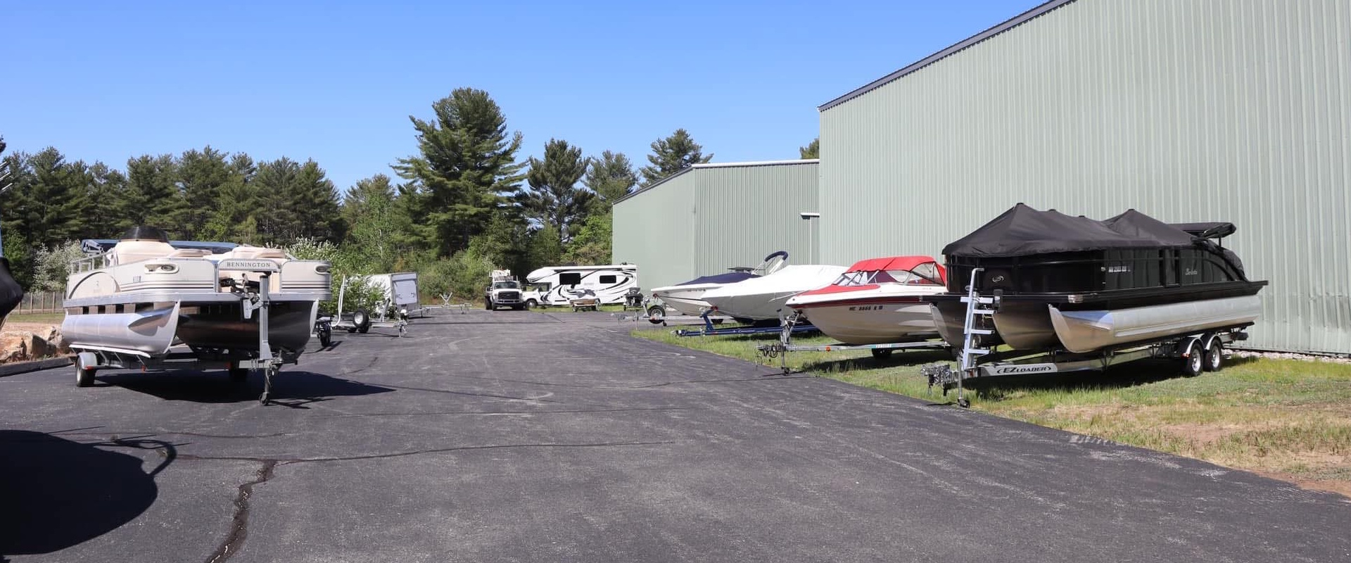 Boats on trailers outside the sage-green Shapleigh boatyard building, with pine trees behind